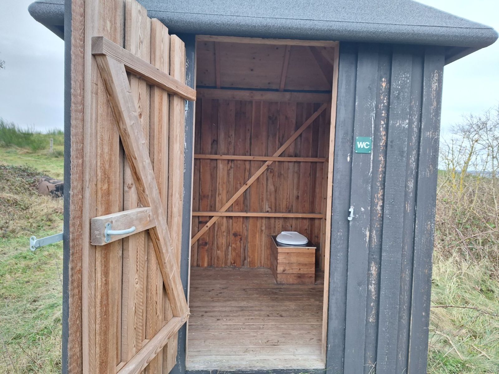 Toilettet er med et stalddørsgreb og åbner udad. Toilettet er mærket med et lille wc skilt til højre for døren. 
