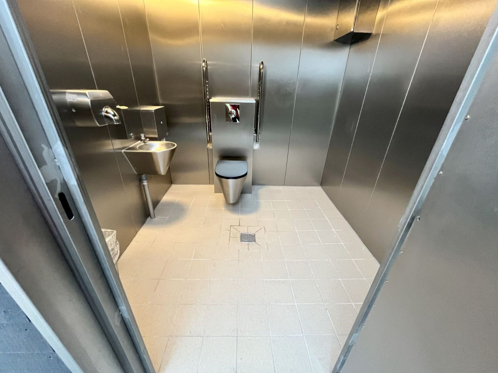 Toilettet med god plads og armstøtter, håndvask, toiletpapir, sæbedispenser, skraldespand og håndtørrer.