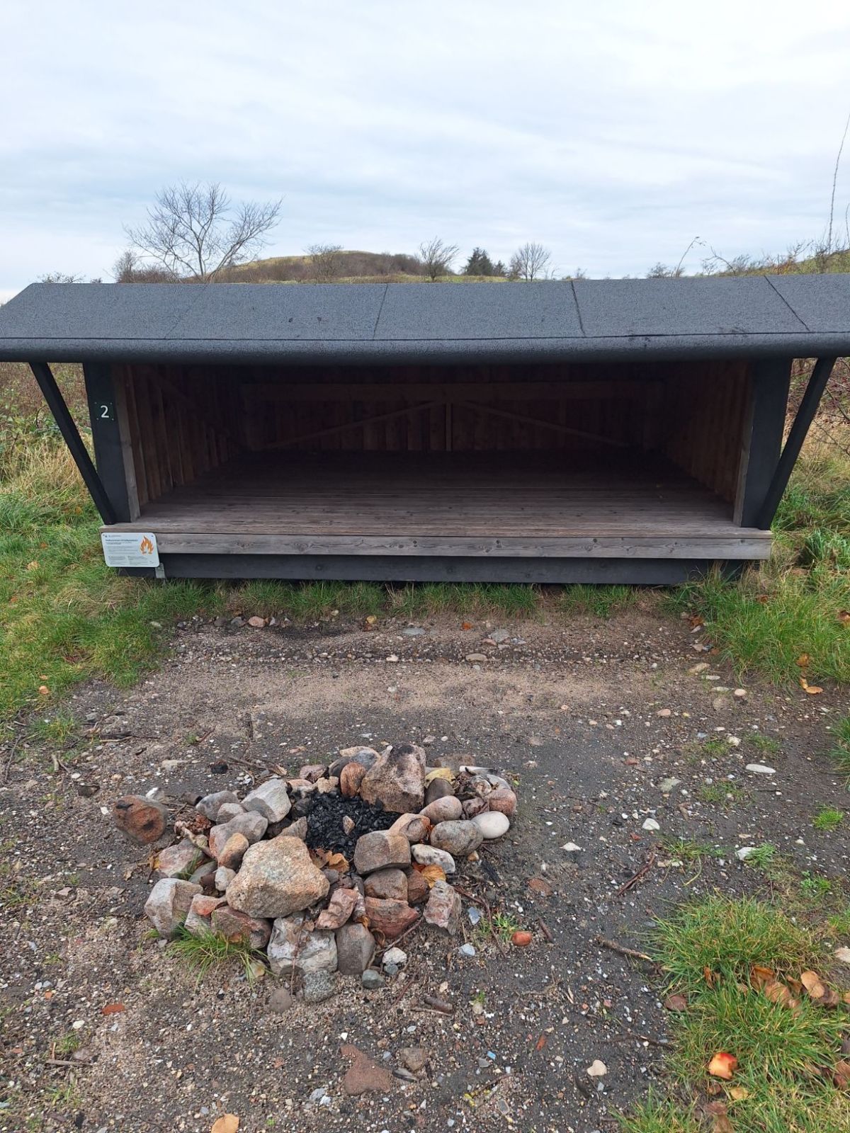 Bålplads foran shelter nummer 2. På shelteren til venstre hænger infotavle om brug af åben ild og forholdsregler herfor. Bålpladsen er med sandet underlag og det øvrige areal er med græstæppe.