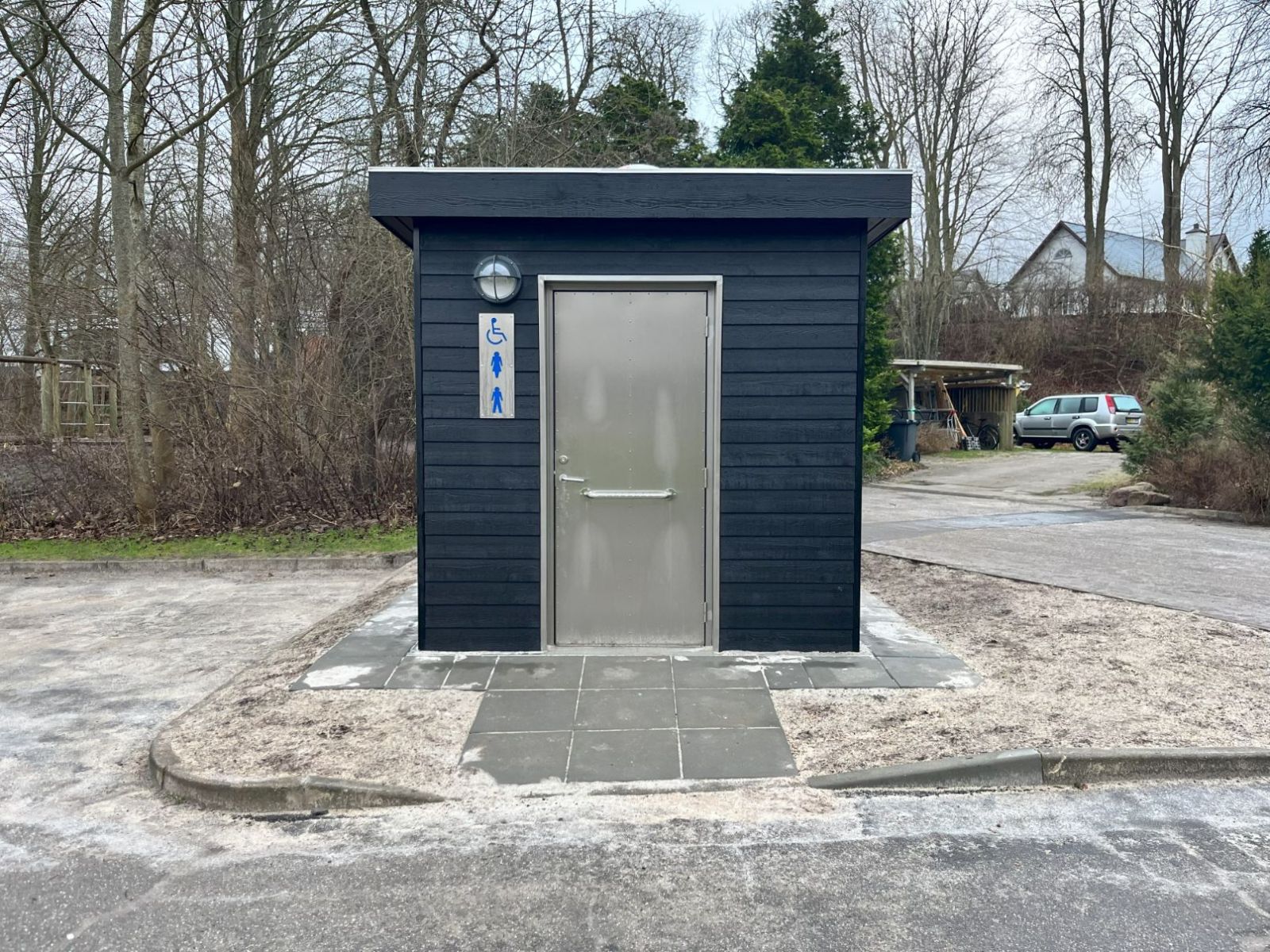 Sort toiletbygning med flisebelagt adgangssti med svag hældning fra asfaltvejen