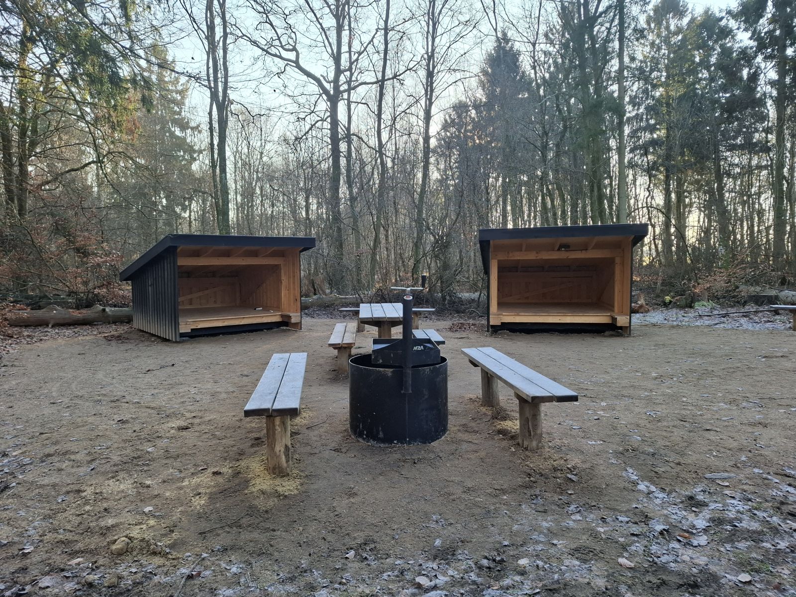 Billedet viser et bålsted med to bænke, grillrist og forhøjet bålring. Bag dette ses et bordebænkesæt, hvis ene bænk er halveret og bag dette ligger to shelters med indgangen pegende mod kameraet. 