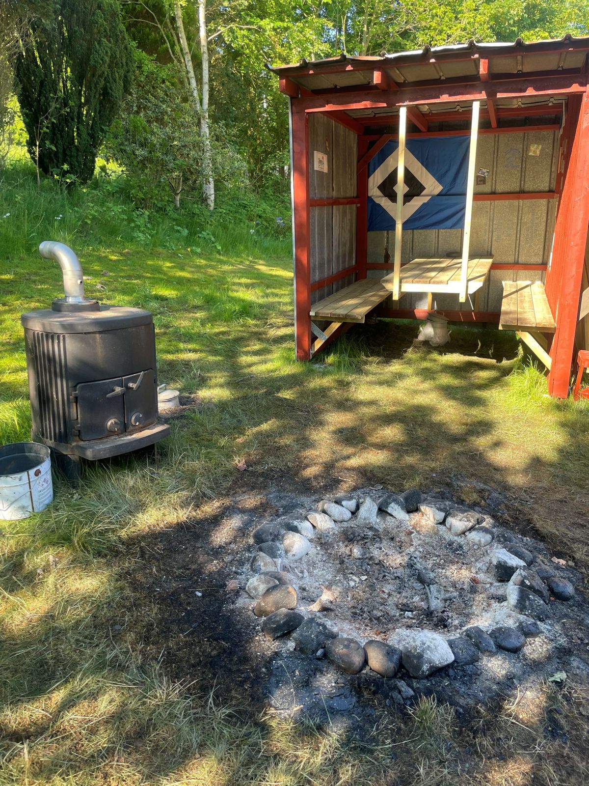Her ses bålstedet med brændeovn og overdækket bordbænkesæt i baggrunden 