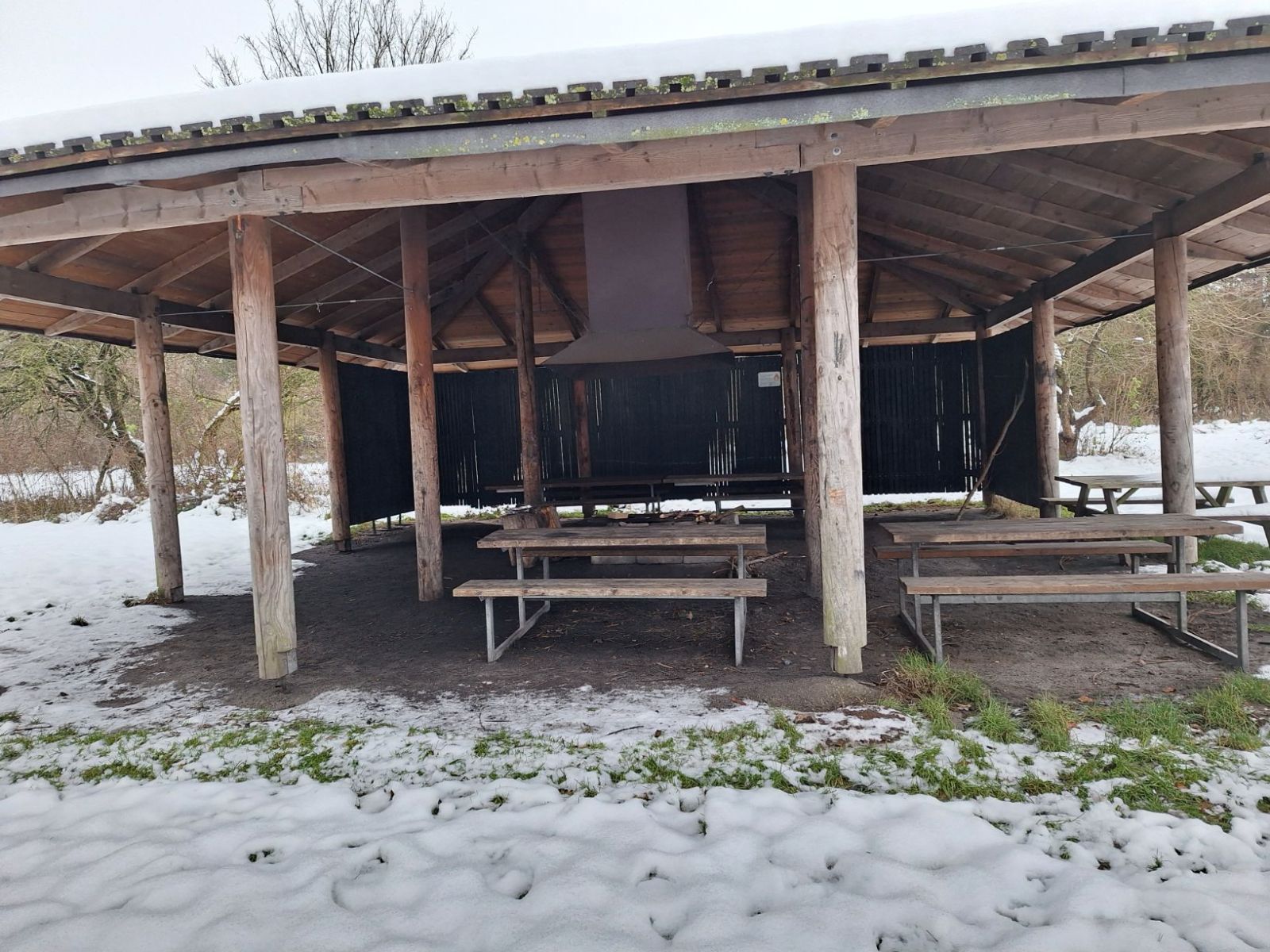 Billede af bålhytten set fra tarzanbanen. Tre almindelige bordebænkesæt samt to kørestolsvenlige bordebænkesæt bagerst i hytten. Bålstedet med skorsten i midten af hytten.