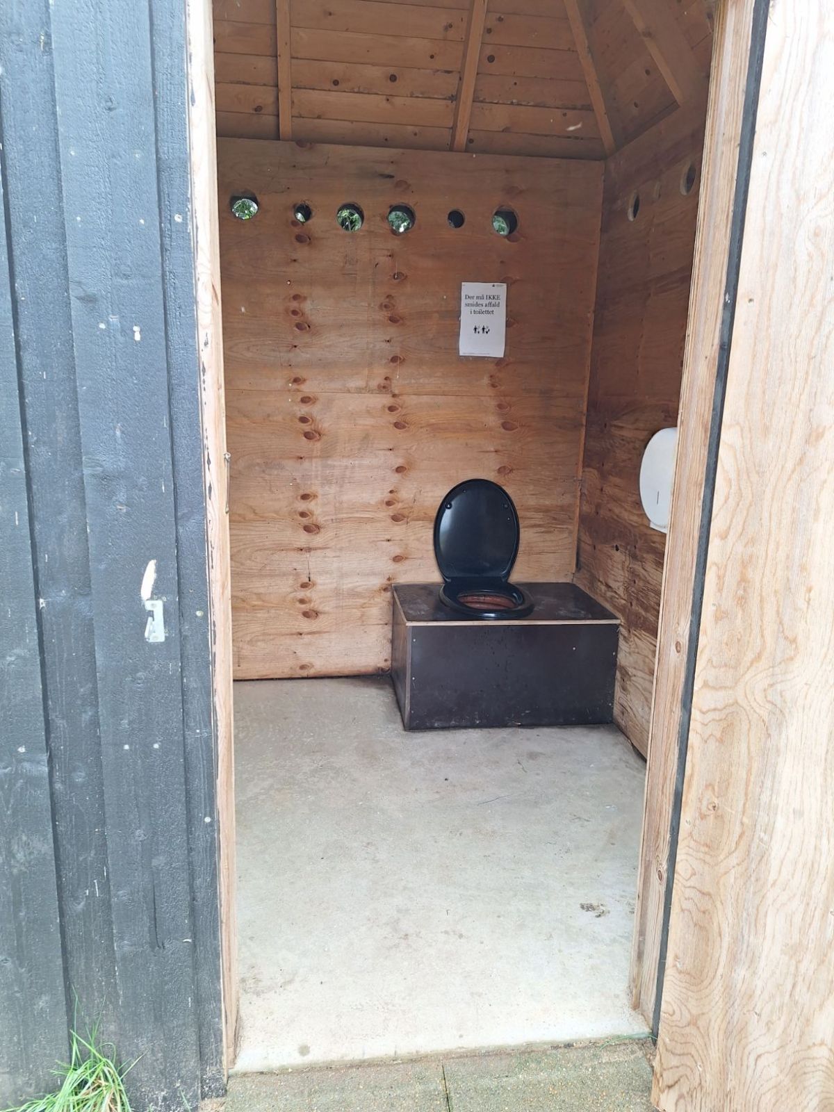 Multtoilettet med døren åbent. Toilettet er placeret til højre for indgangen med frirum til venstre på godt 85 centimeter. Fra døren til toilettet er der 130 centimeter frirum. Toiletpapiret er monteret på væggen til venstre for brugeren. Fra fliserne fo