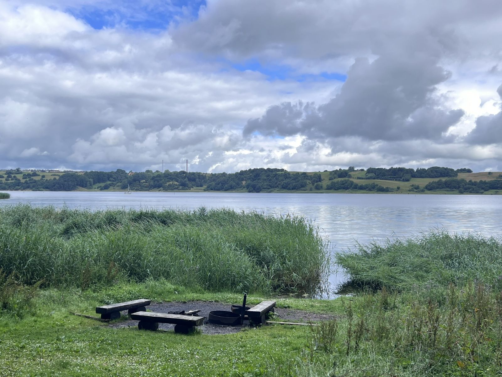 Bålplads med tre bænke uden ryglæn. Bålpladsen ligger lige ved vandkanten af Mariager Fjord. I baggrunden ses fjordskrænte på modsatte side. Vatskyer på himlen.