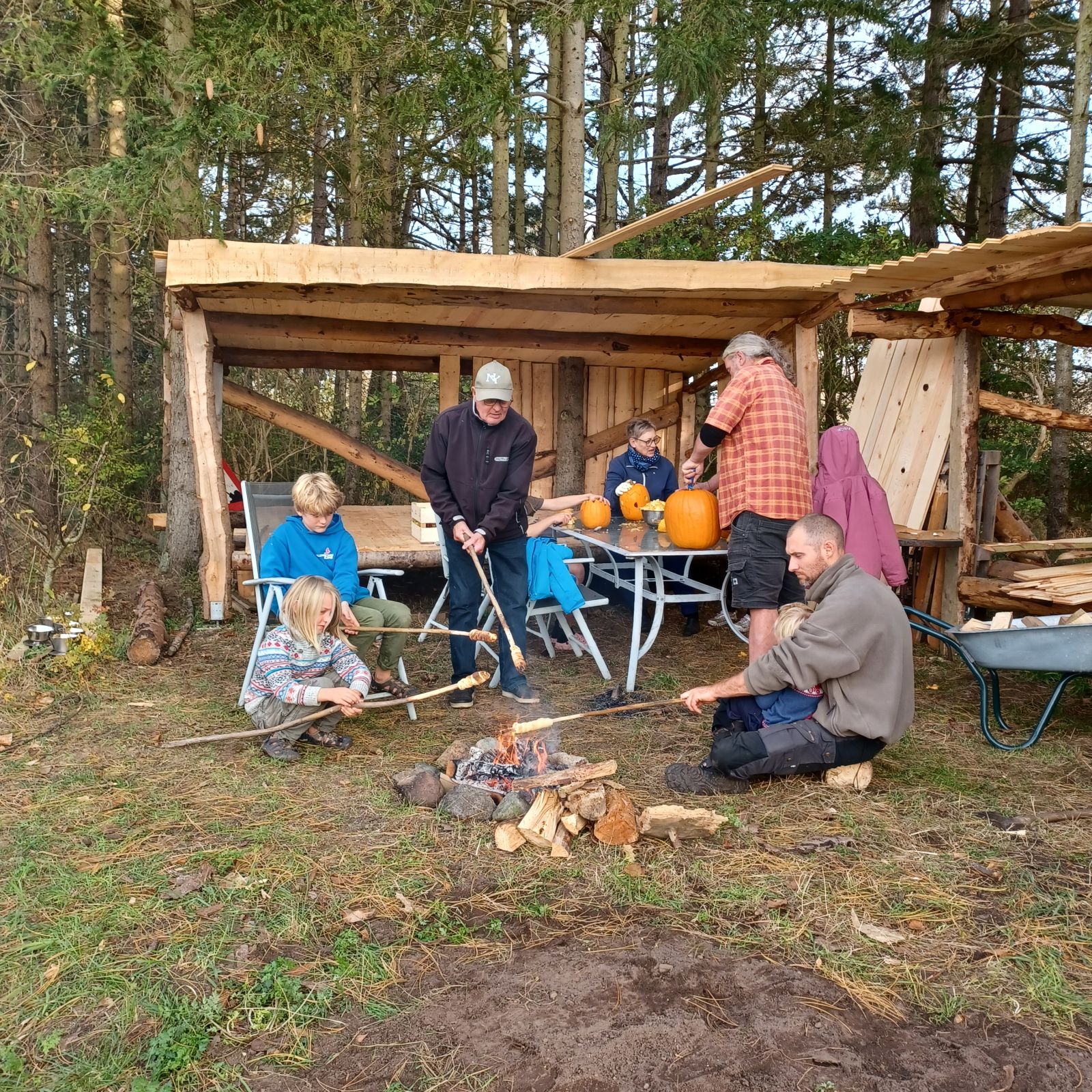 En gruppe mennesker, både børn og voksne, laver snobrød over et bål nær et træshelter; i baggrunden står et bord med græskar og redskaber
