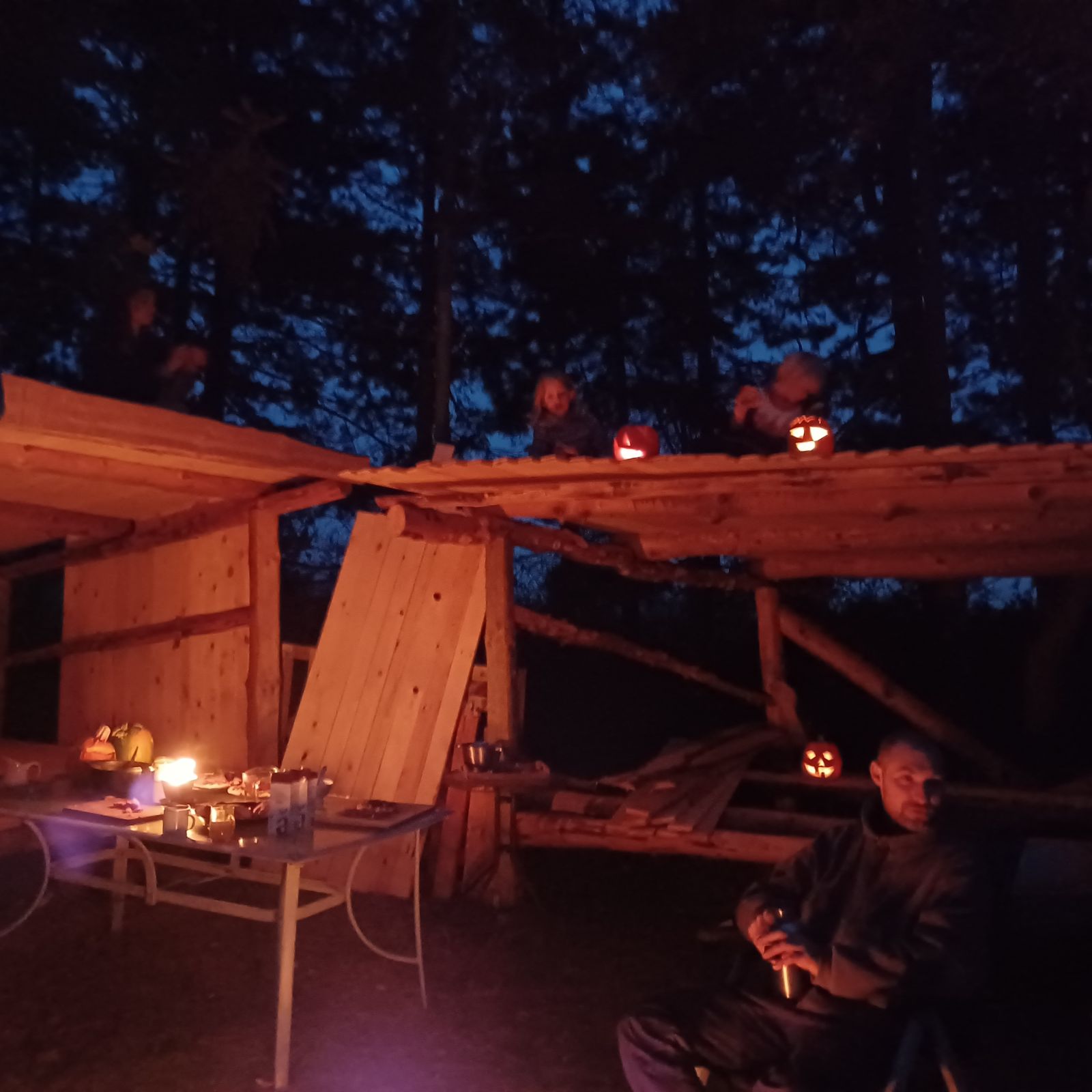 En gruppe mennesker, både børn og voksne sidder ved de to shelters om natten. Der er lyst op af stearinlys.