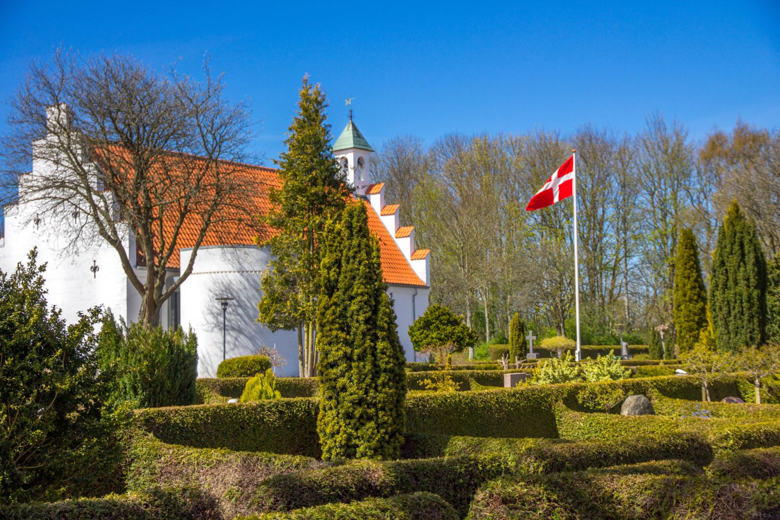 Hvidkalket kirke med rødt tegltag i venstre side af billedet. Kirkegår med fint-klippede lave hække i forgrunden og flagstang med hejst Dannebrogsflag