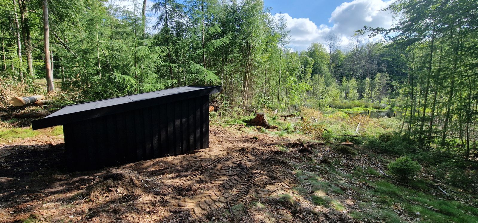 Billedet viser et lille, sortmalet shelter bagfra. Shelteret står i en lysning i skoven og indgangen peger ud mod en mose med åbent vandspejl. Omkring pladsen vokser nåletræer og lidt løvtræer i forskellige højder. Terrænet er let kuperet.