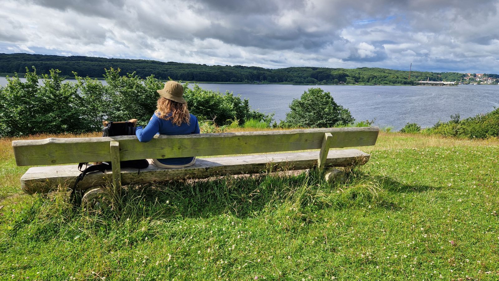 Lang bænk i massiv bænk placeret på toppen af en bakke med udsigt udover Mariager Fjord. Hobro anes i det fjerne i højre hjørne.