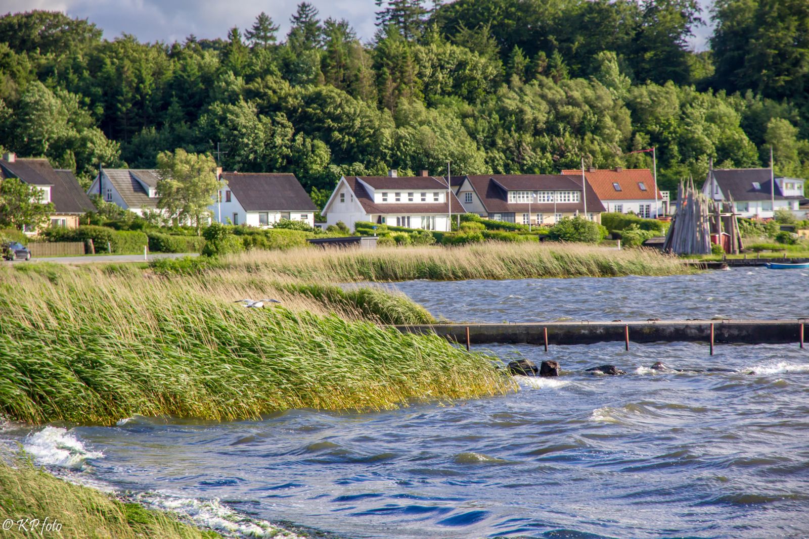 Huse ses i baggrunden. Husene ligger ud til kysten, som ses forrest i billedet. Der er små bølger i vandet og en bro strækker sig ud mod højre i billedet.
