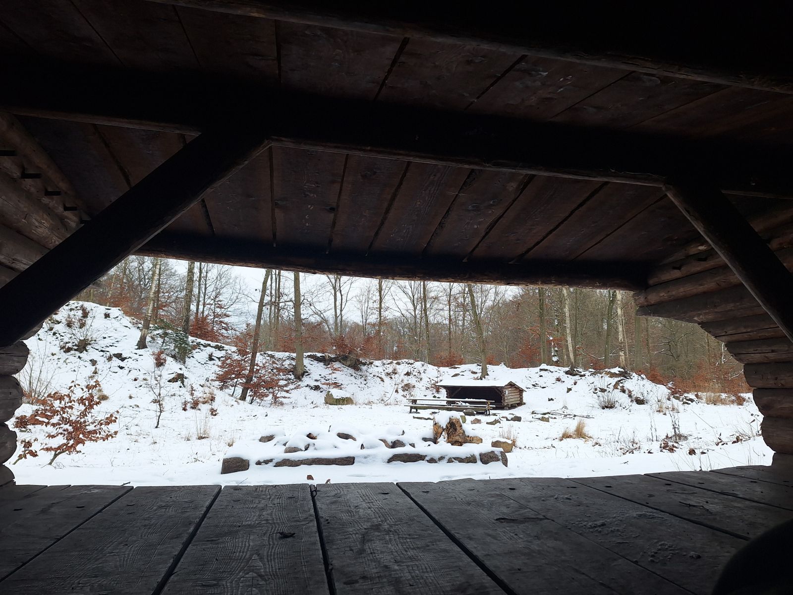 Billedet viser udsigten fra indgangen til et shelter. Her ses et bålsted omgivet af sten i en firkantet formation. Længere væk ses et andet shelter, samt bordebænkesæt og endnu et bålsted. Der ligger sne.