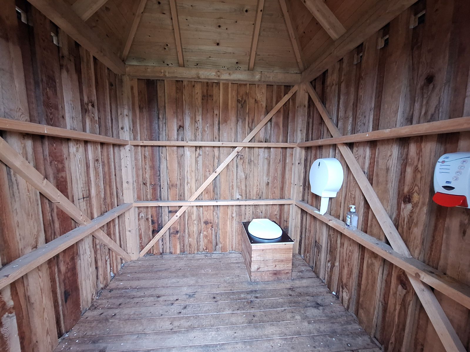 Multtoilettet er placeret i højre hjørne for døråbningen og med vægmonteret toiletpapirsholder samt håndsprit. Gulvet er træbrædder. 