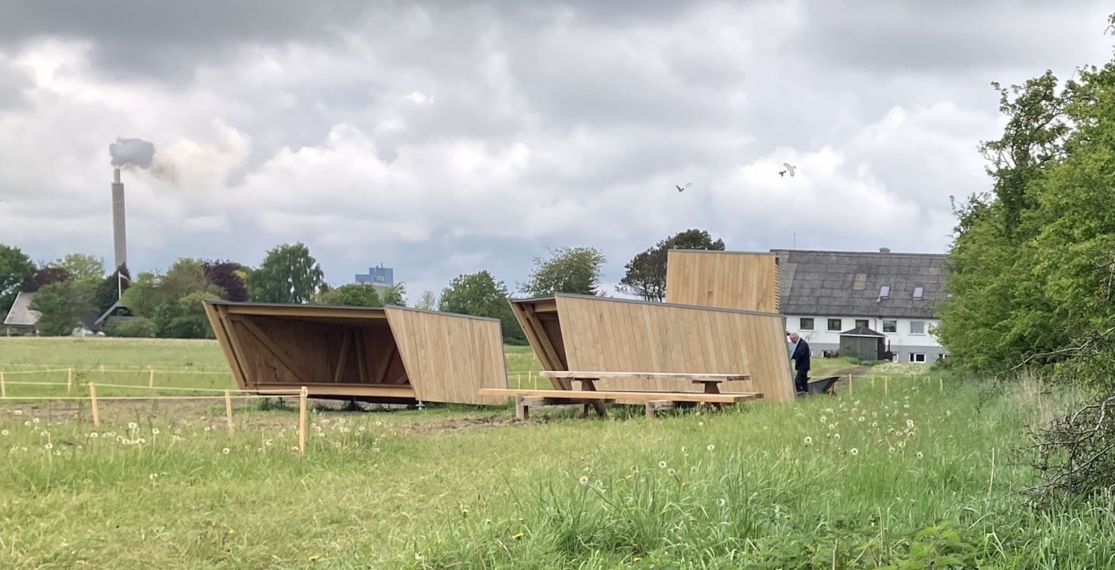 Shelters i ubehandlet træ på åben plads med huse i baggrunden