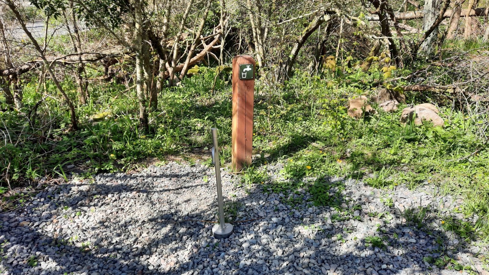 En stålvandpost på et underlag af grove skærver. I baggrunden ses en rød pæl med symbol for drikkevand på.