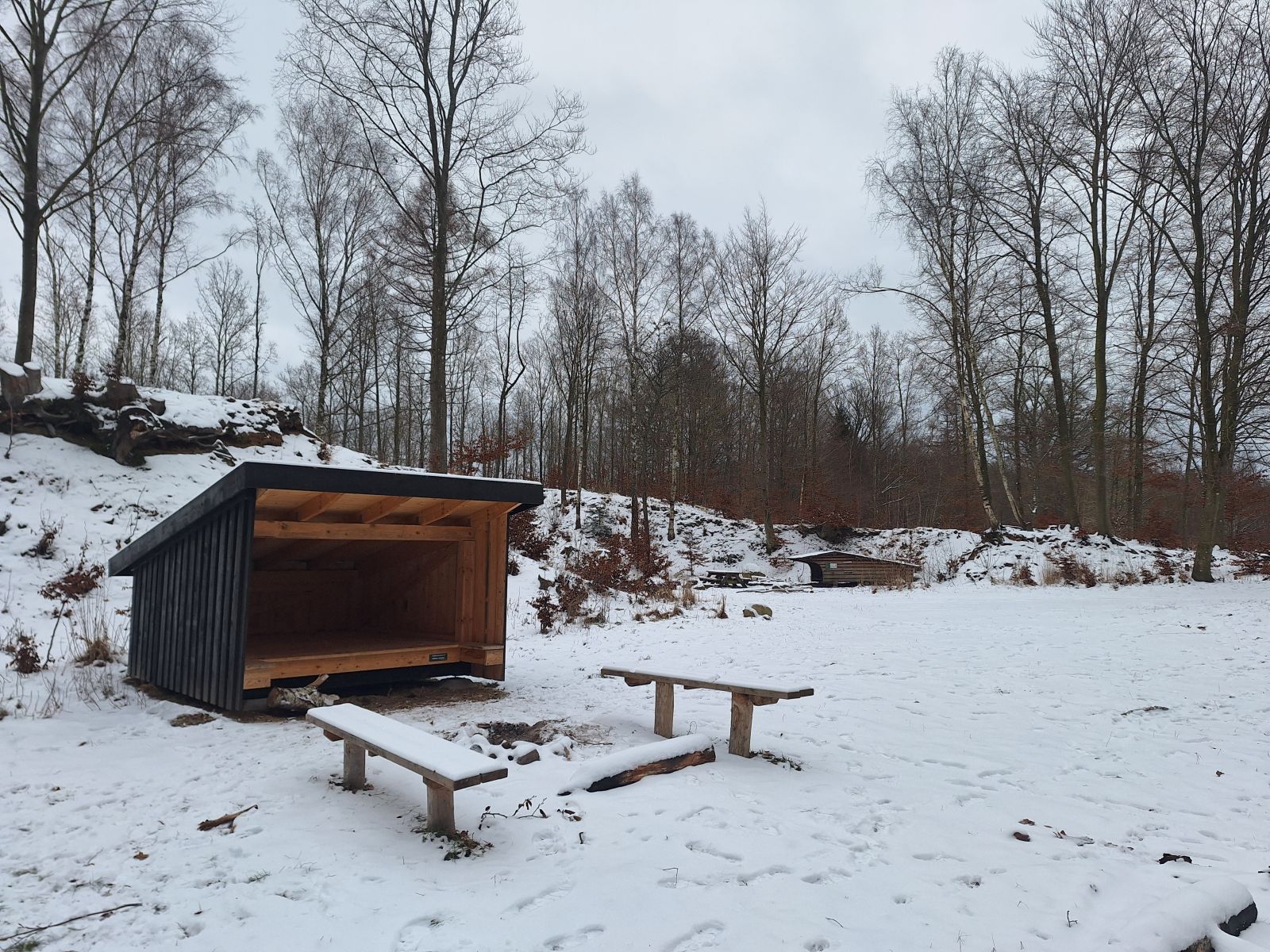 Billedet viser et mindre shelter i et sneklædt landskab. Foran shelteret er et bålsted med en bænk på hver side. I baggrunden anes et andet shelter. Løvtræer vokser spredt. 