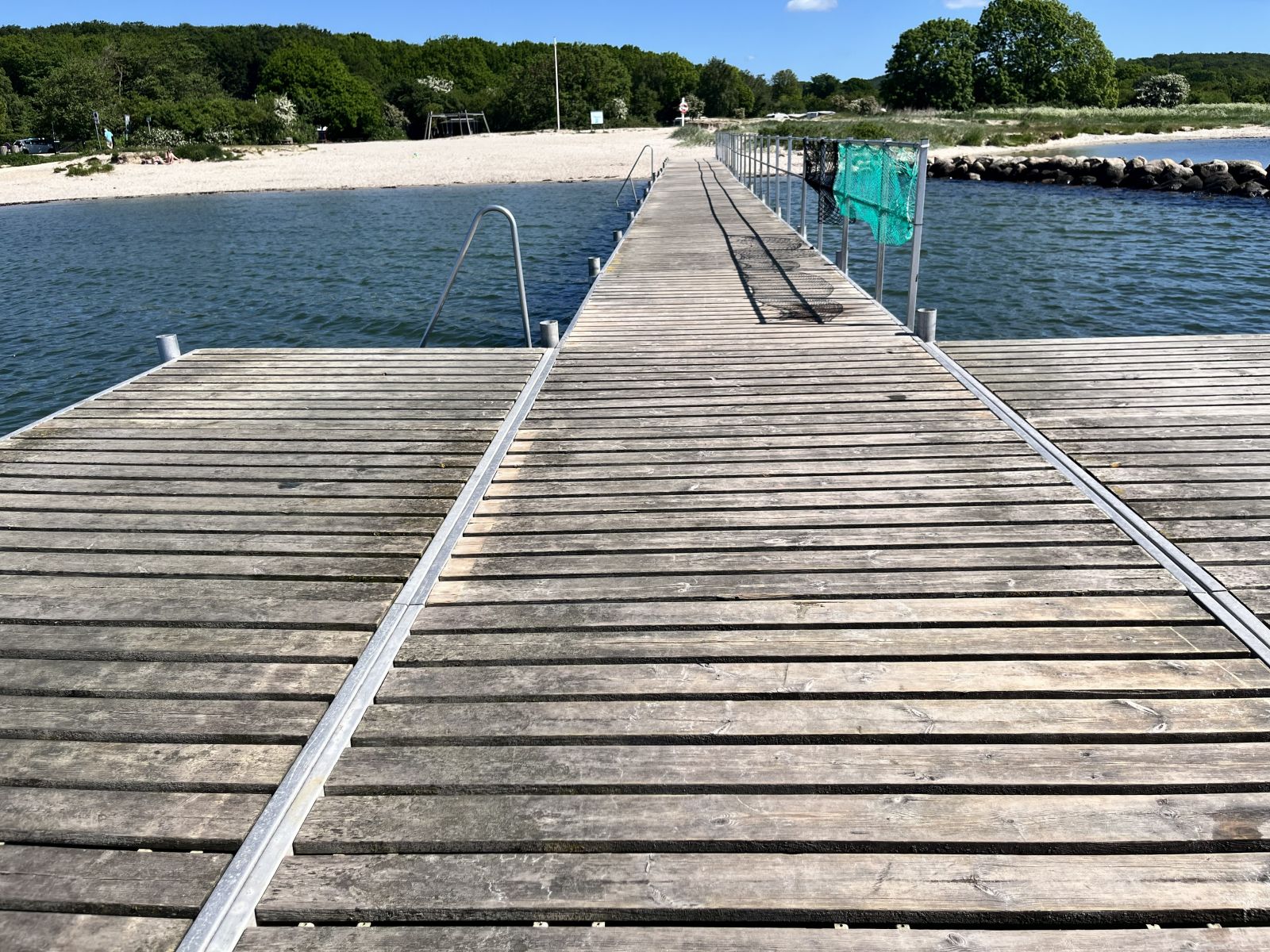 Badebroen med udsigt mod stranden. Der er en platform med 2 stiger helt yderst og langs broen er der gelænder på den ene side
