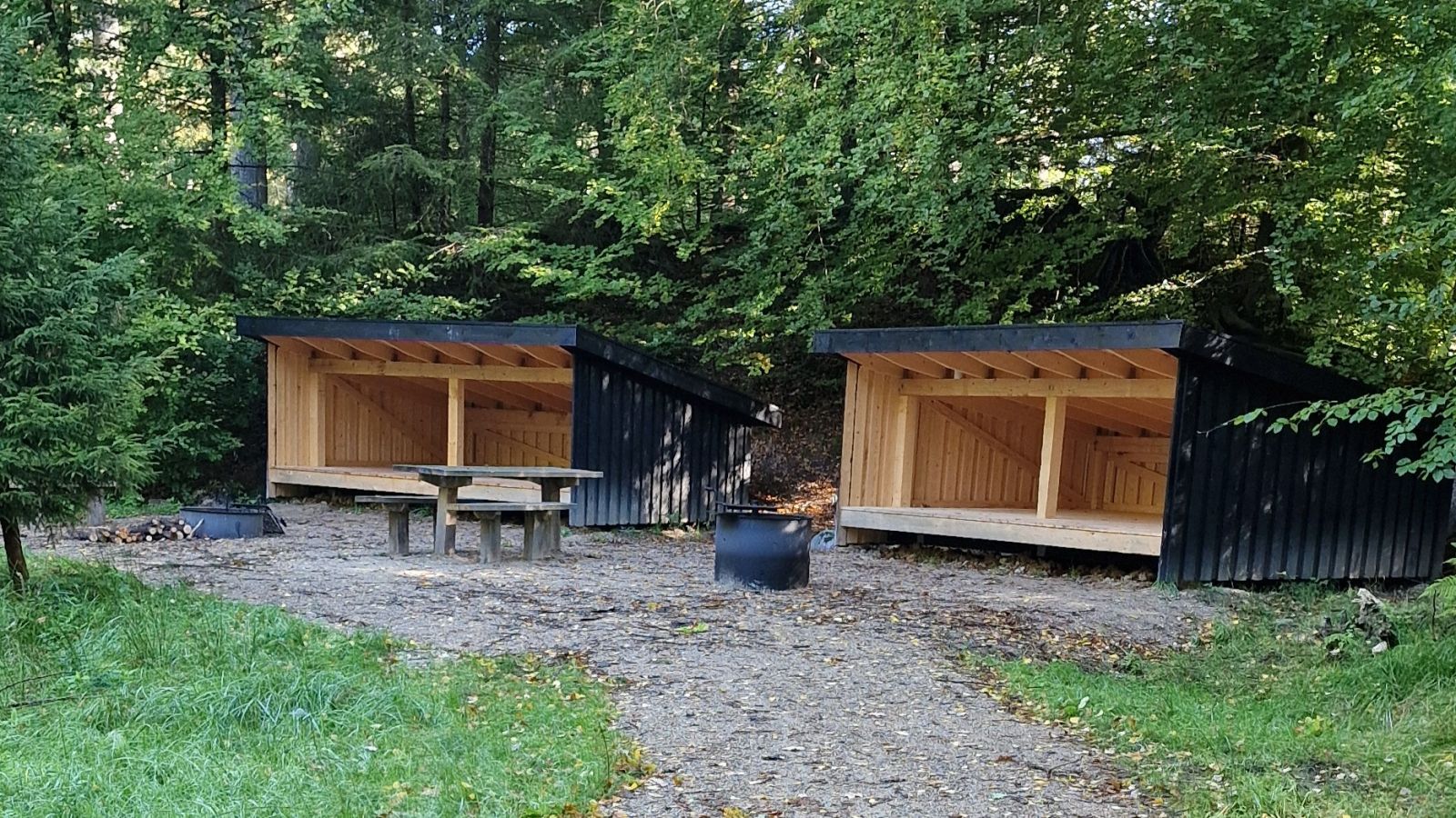 Billedet viser to større, sortmalede sheltere ved siden af hinanden, set forfra. Shelterene er orienteret næsten parallelt med indgangene pegende i samme retning. Foran shelteret til venstre på billedet er et bålsted med en ca. 30 centimeter høj bålring. Foran shelteret til højre er et bålsted med en ca. 60 centimeter høj bålring. Der er monteret grillriste på begge bålsteder. Foran, i midten, står et bord-og-bænkesæt, hvor den ene af bænkene er halv så lang som den anden, så der er plads til at sidde ved bordet med og uden kørestol. Lige bag shelterene starter bøgeskoven. Underlaget er grus.