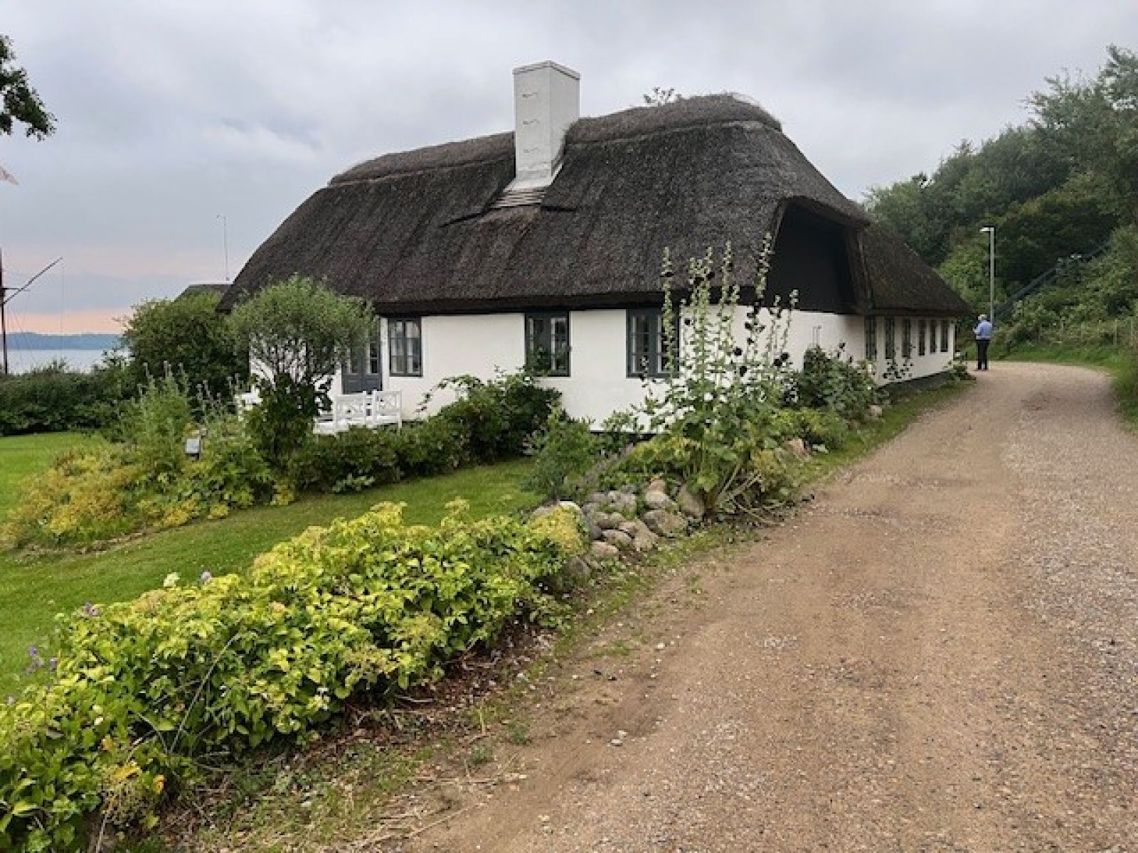 Hvidt stråtækt sommerhus omgivet af en frodig have og en grusvej. I baggrunden ses et vandområde og skov.