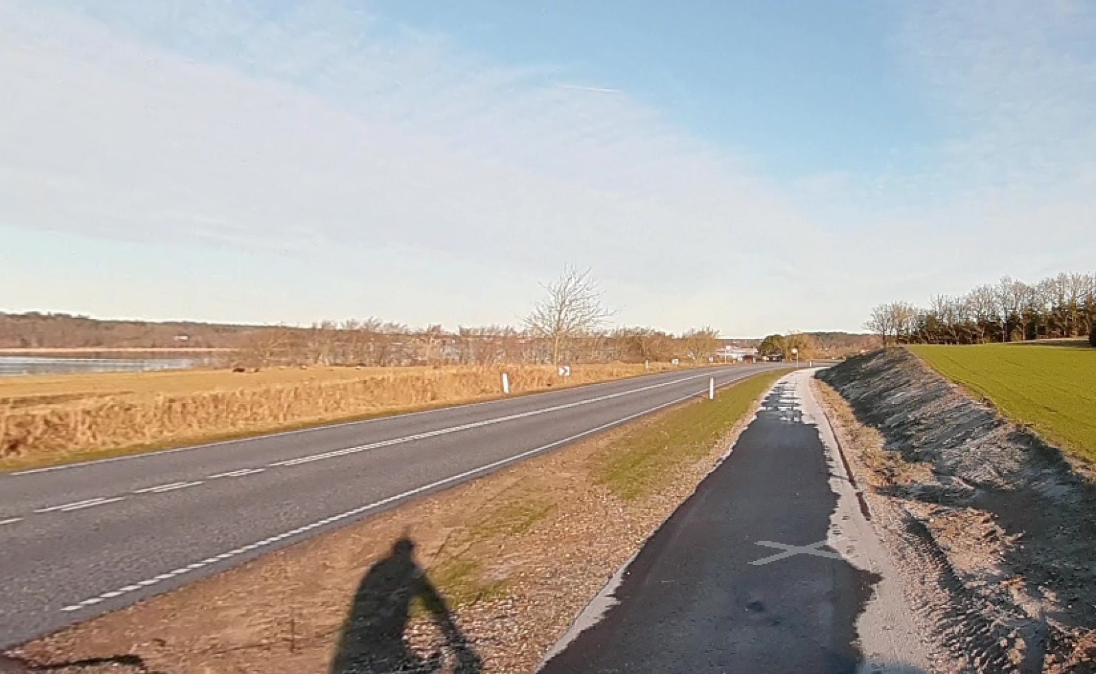 En asfalteret cykelsti langs en landevej i et åbent, bakket landskab med efterårsfarvede træer i baggrunden.