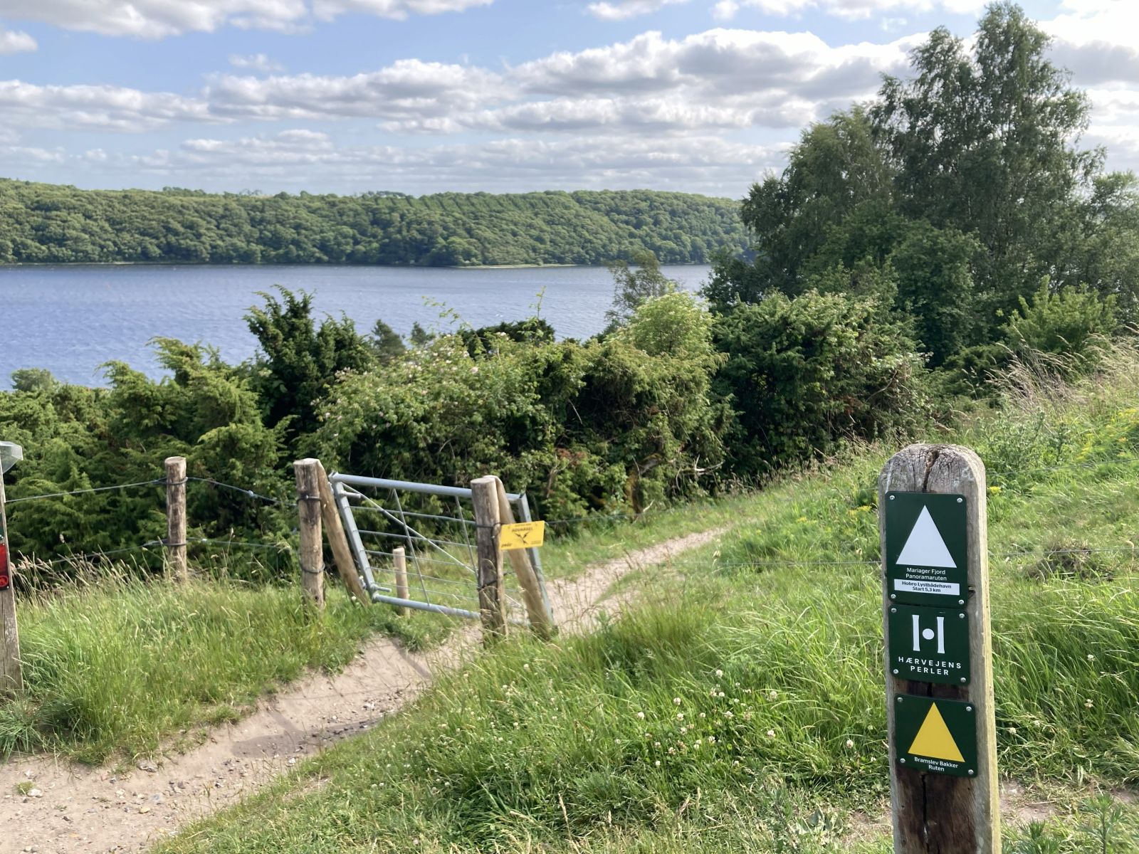 Udsigt over Mariager Fjord. Markeringspæl i højre side med en hvid og en gul trekant pegende opad. Klaglåge henover stien.