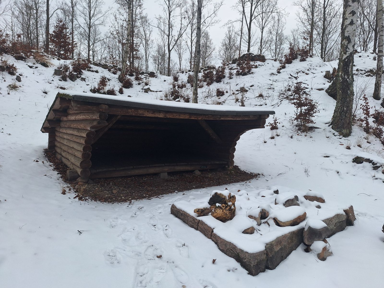 Billedet viser et shelter bygget af bjælker. Foran shelteret er et bålsted omgivet af sten i en firkantet formation. I baggrunden ses lidt birketræer. Der ligger sne.