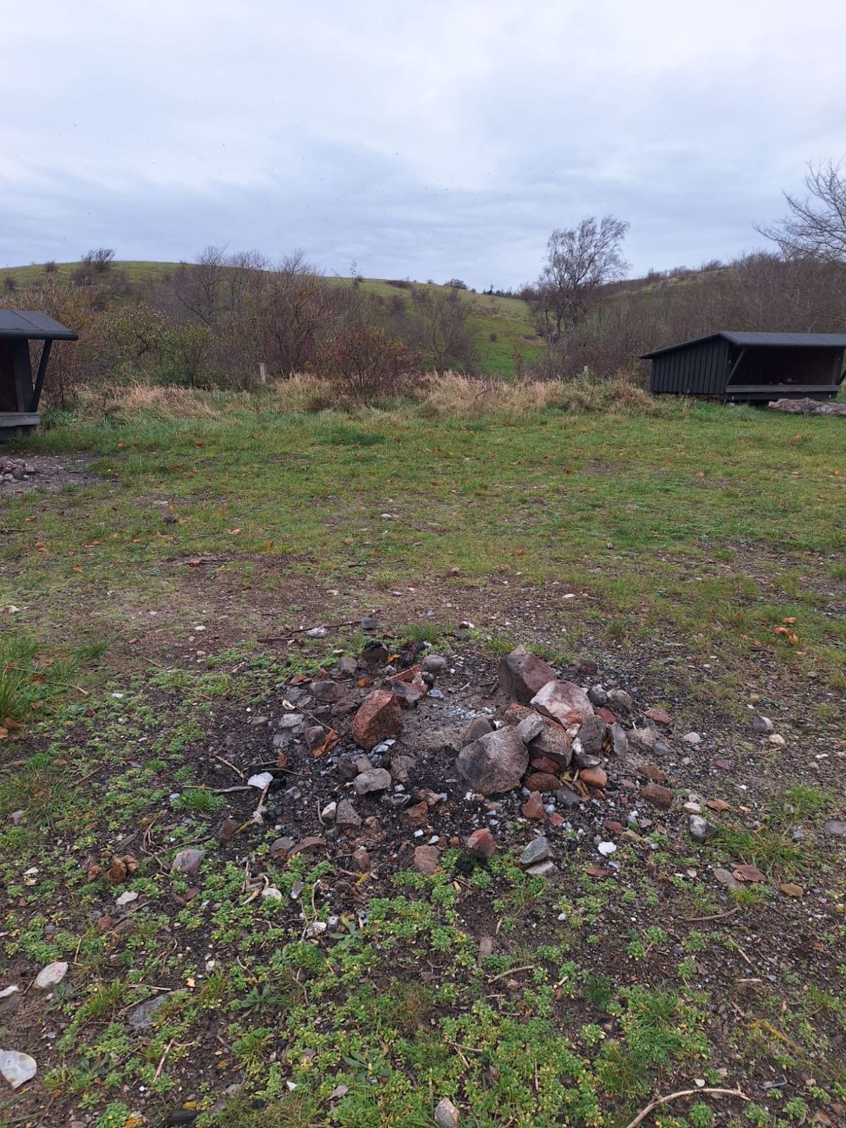 Bålplads midt på pladsen med shelter 2 og 3 i baggrunden. Græstæppe omkring bålpladsen som er afgrænset med sten og jord i bunden.