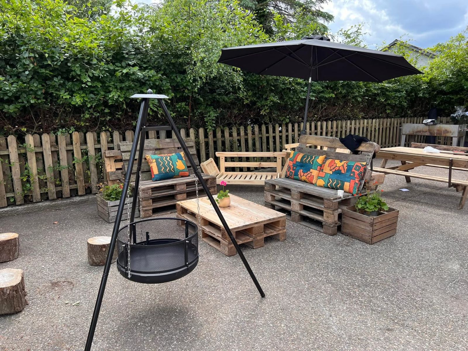 Et hyggeligt udendørd opholdsområde indrettet med pallemøbler og et tripodbålsted med hængegrill.