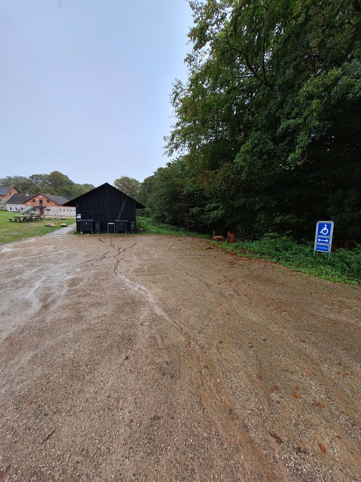 Gavlen af Hald Laden set fra handicapparkeringen. Sti med fast belægning af stampet grus leder venstre om bygningen til toiletterne. Sti mod højre leder ned til naturlegepladsen. 