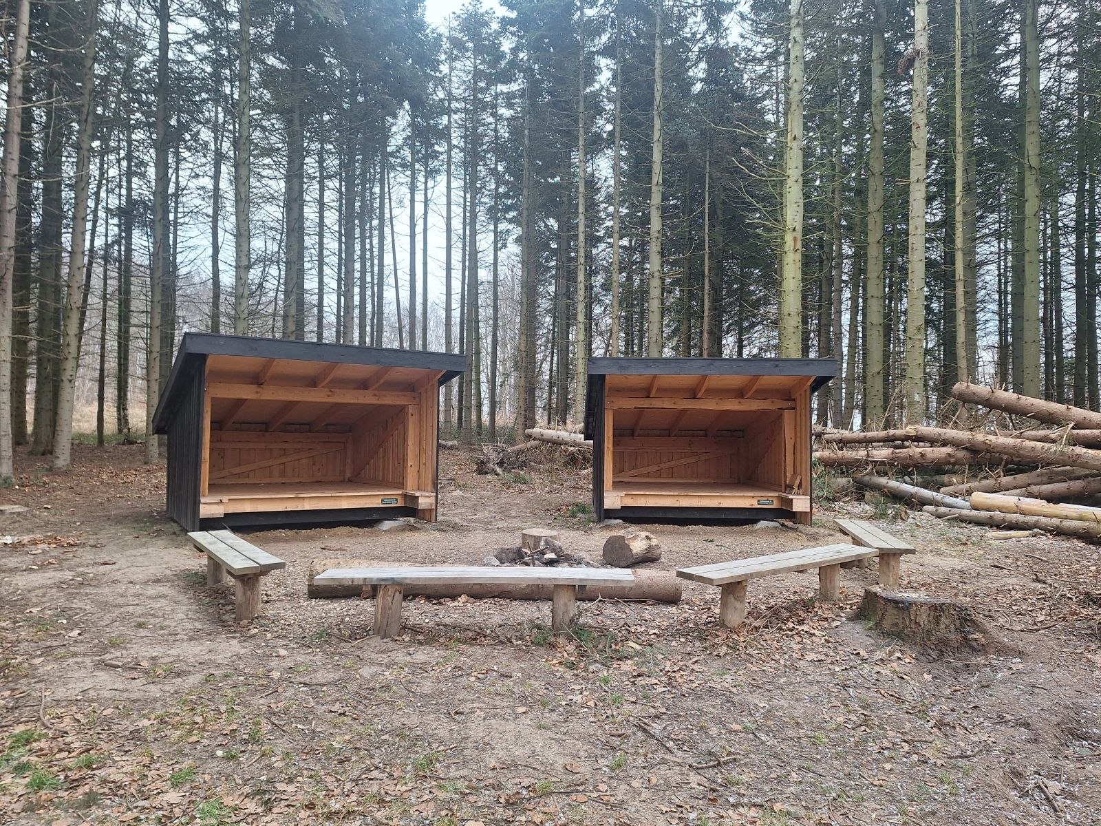 Billedet viser to shelters og et foranliggende bålsted omgivet af fire bænke. I baggrunden står høje grantræer.