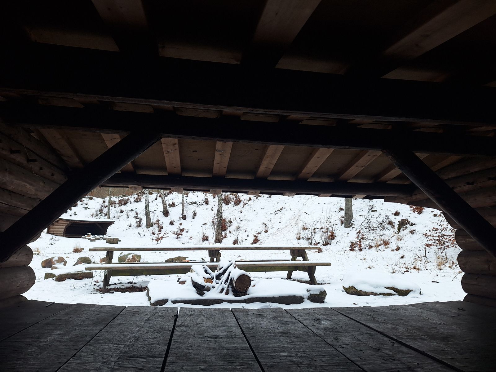 Billedet viser udsigten fra en shelterindgang. Nærmest ses et bålsted, herefter et bordebænkesæt med plads til ca. 12 personer. I baggrunden ses et andet shelter, ca. 30 meter væk, med indgangen pegende mod kameraet. 