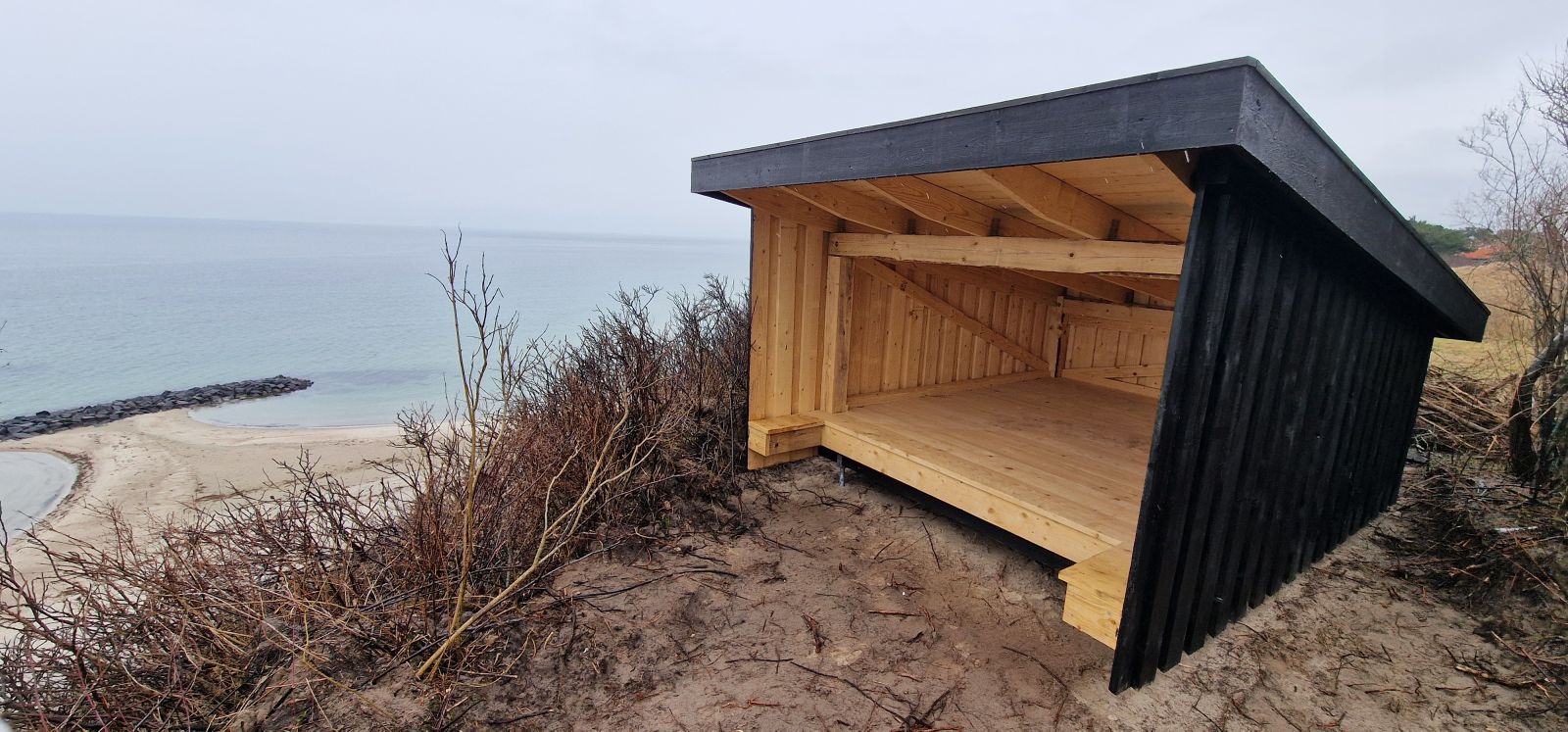 Billedet viser et lille, sortmalet shelter beliggende på toppen af en skrænt. Underlaget er fast sand. I baggrunden ses strandkanten og havet.