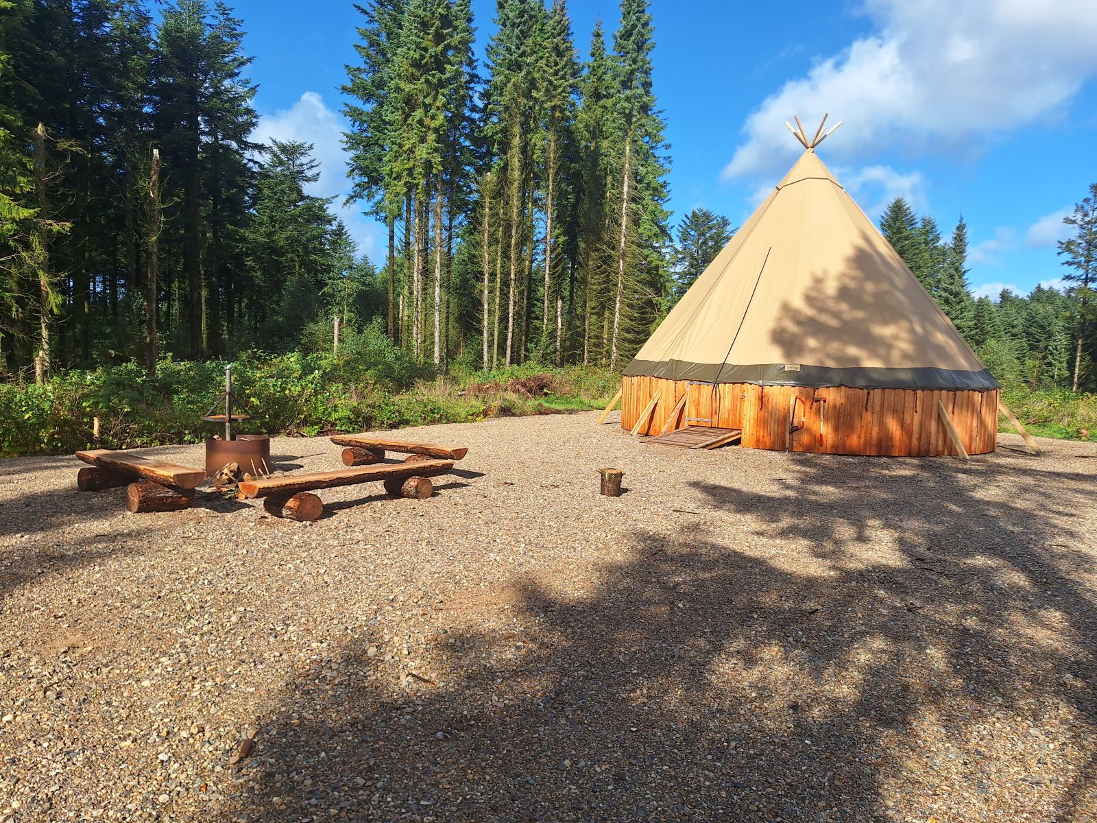 Billedet viser teltet som har form som en tipi, bålpladsen ude foran og skoven lige bagved. Underlaget er grus.