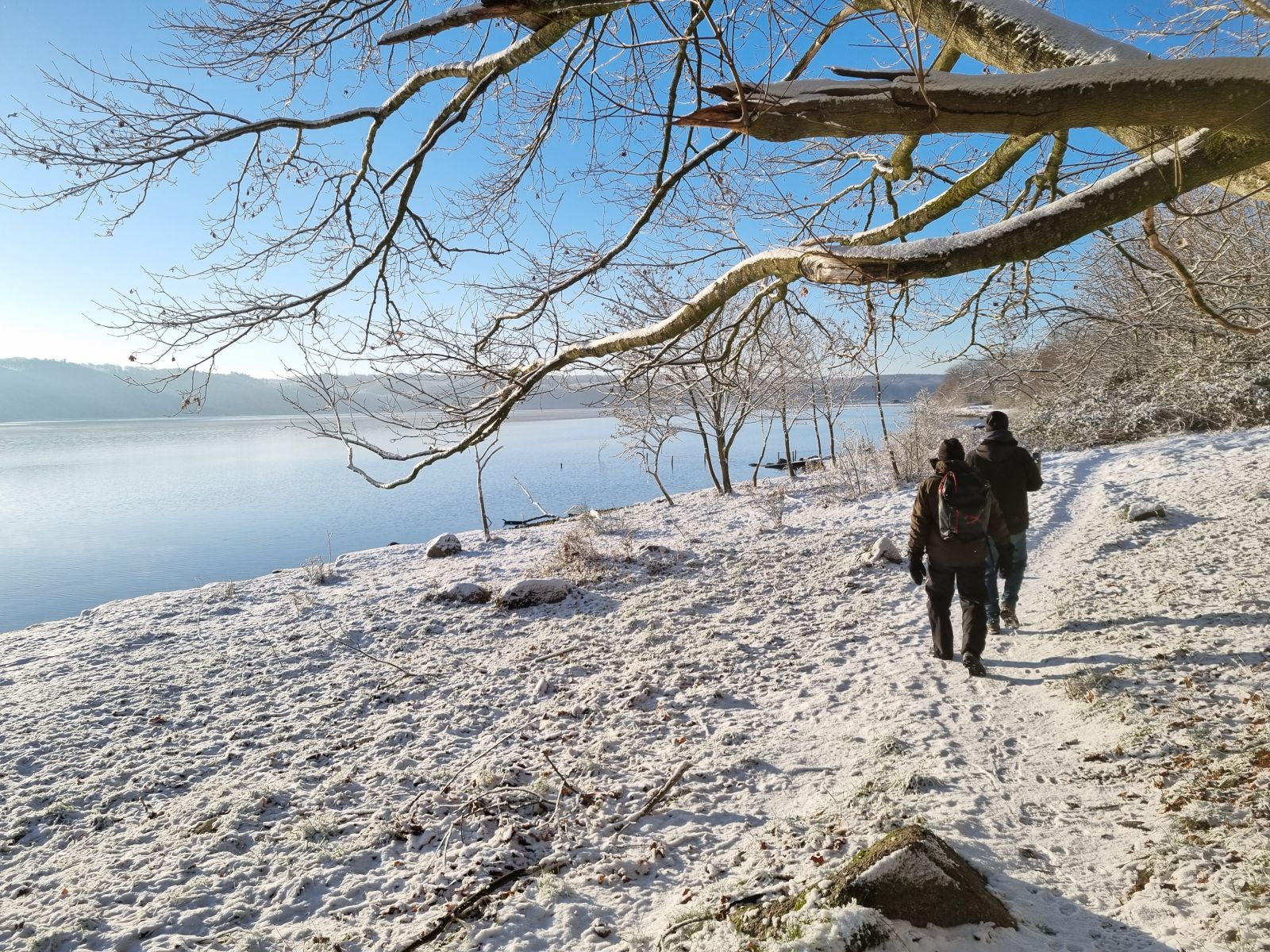 Snedækket vinterlandskab med klar blå himmel og en stille Mariager Fjord til venstre i billedet. To vandrere går væk fra kameraet i højre side.