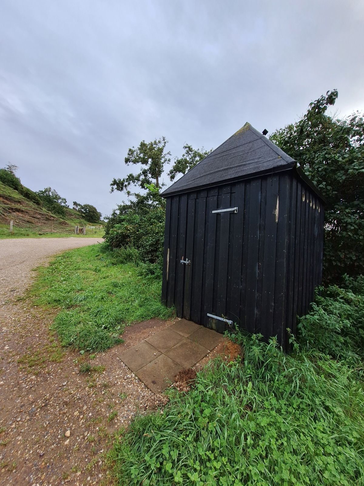 Multtoilettet set med indgangen til parkeringspladsen i baggrunden. Toilettet samt den flisebelagte sti er med vegetation omkring sig. 