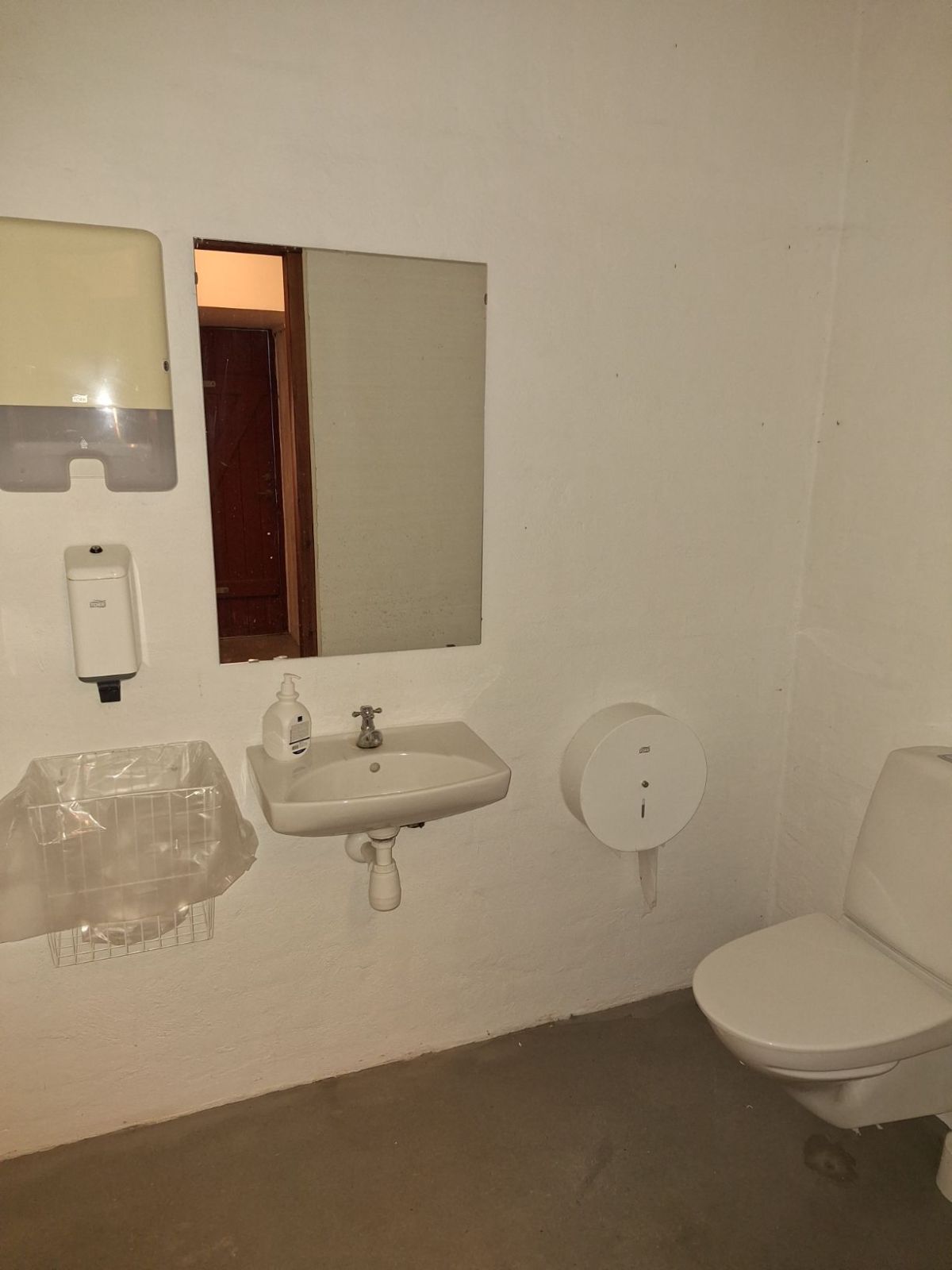 Det første toilet for indgangen med toilettet til højre og væghængt toiletpapirsholder, håndvask med spejl, skraldespand og sæbedispenser og papirshåndklædeholder på bagerste væk