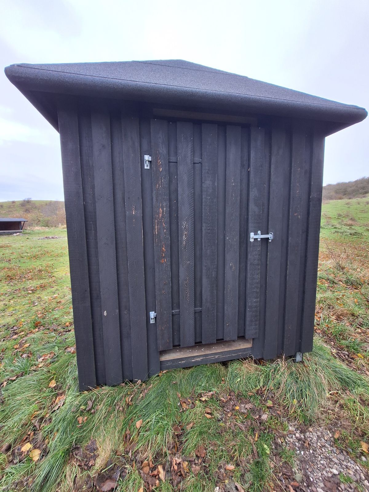 Multtoilettet set udefra med shelter 3 i baggrunden. Underlaget er græstæppe foran bygningen og med et trin på cirka 13 centimeter ind til toilettet. 