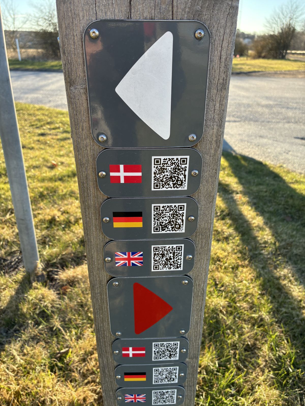 Afmærkningspæl med fire piktogrammer. Øverst en hvid pil pegende mod venstre. De tre nederster er mindre med hver deres QR kode og et flag foran. Øverst dansk, midderst tysk og nederst engelsk.