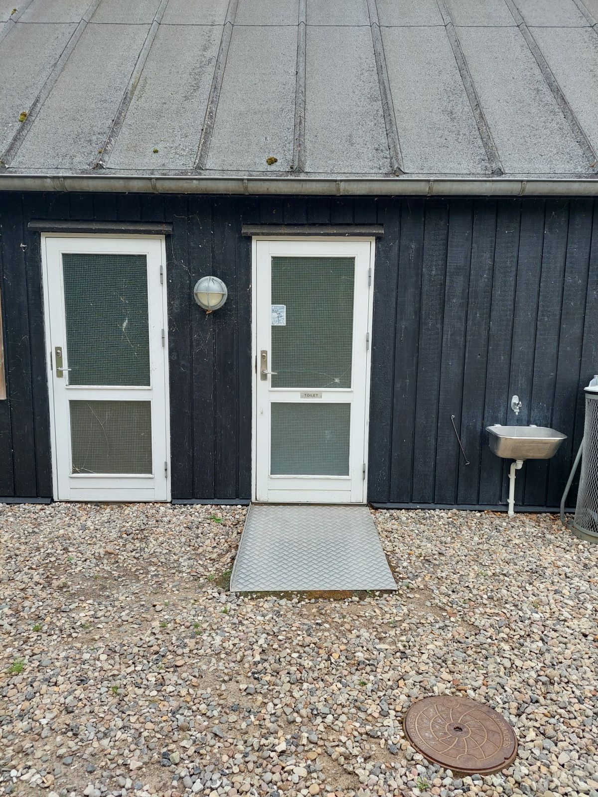 En metalrampe ligger foran døren ind til toilettet. Metalrampen ligger fladt oven på perlegruset. 