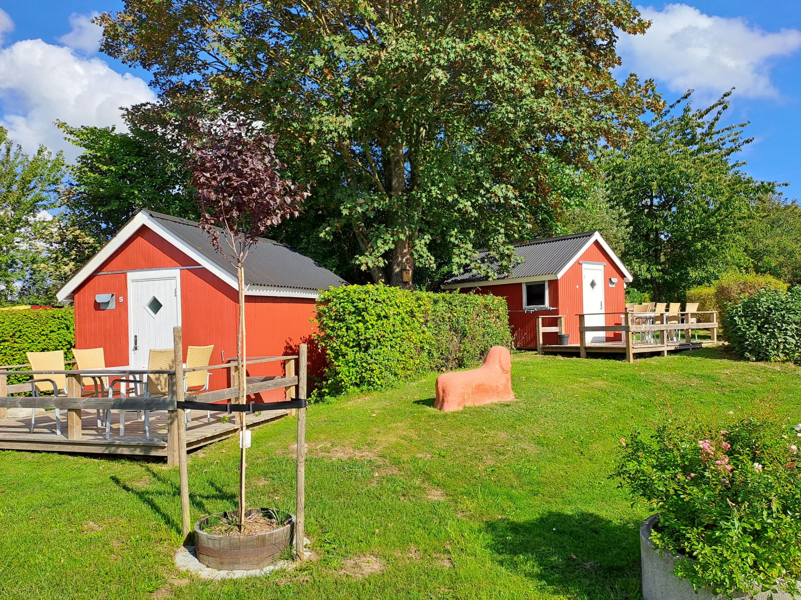 To små røde træhytter med hvide døre og terrasser, omgivet af grønne hække og træer i en solrig have. Der står havemøbler på terrasserne og en rød stenfigur på græsset foran.