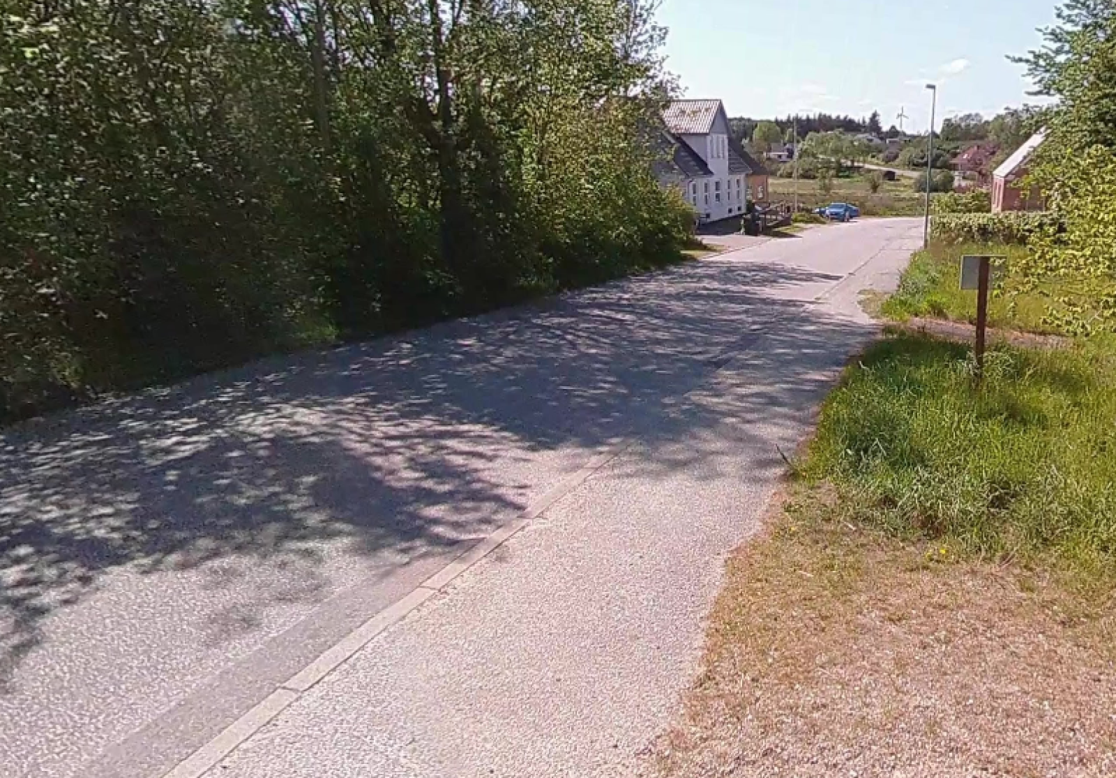 Asfalteret fortov forløber langs vej med træer på modsatte side af vejen og et par huse længere nede.