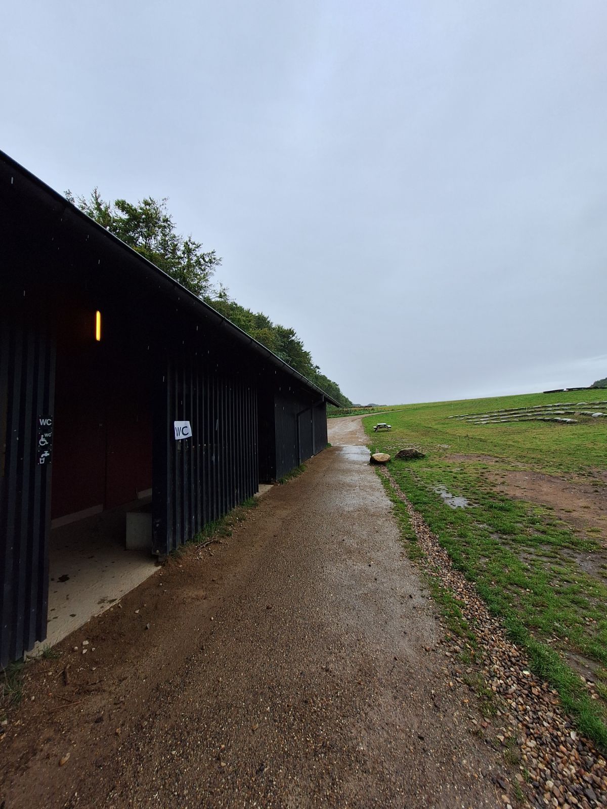 Indgangen til toiletterne ses fra Haldladens Udstilling mod handicap parkeringen. På facaden hænger skilte for toilet, handicap toilet samt drikkevand. Underlaget er jævnt og går fra stampet grus på stien til cement belægning. Græsplæne mod højre.