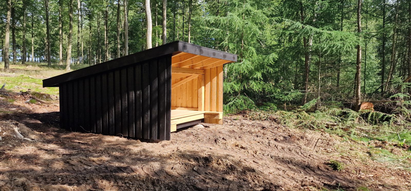 Billedet viser et lille, sortmalet shelter skråt forfra. Shelteret står i en lysning i skoven. Bag shelteret ses nåletræer. Terrænet er let skrånende. Underlaget er delvist græs og delvist bar jord.