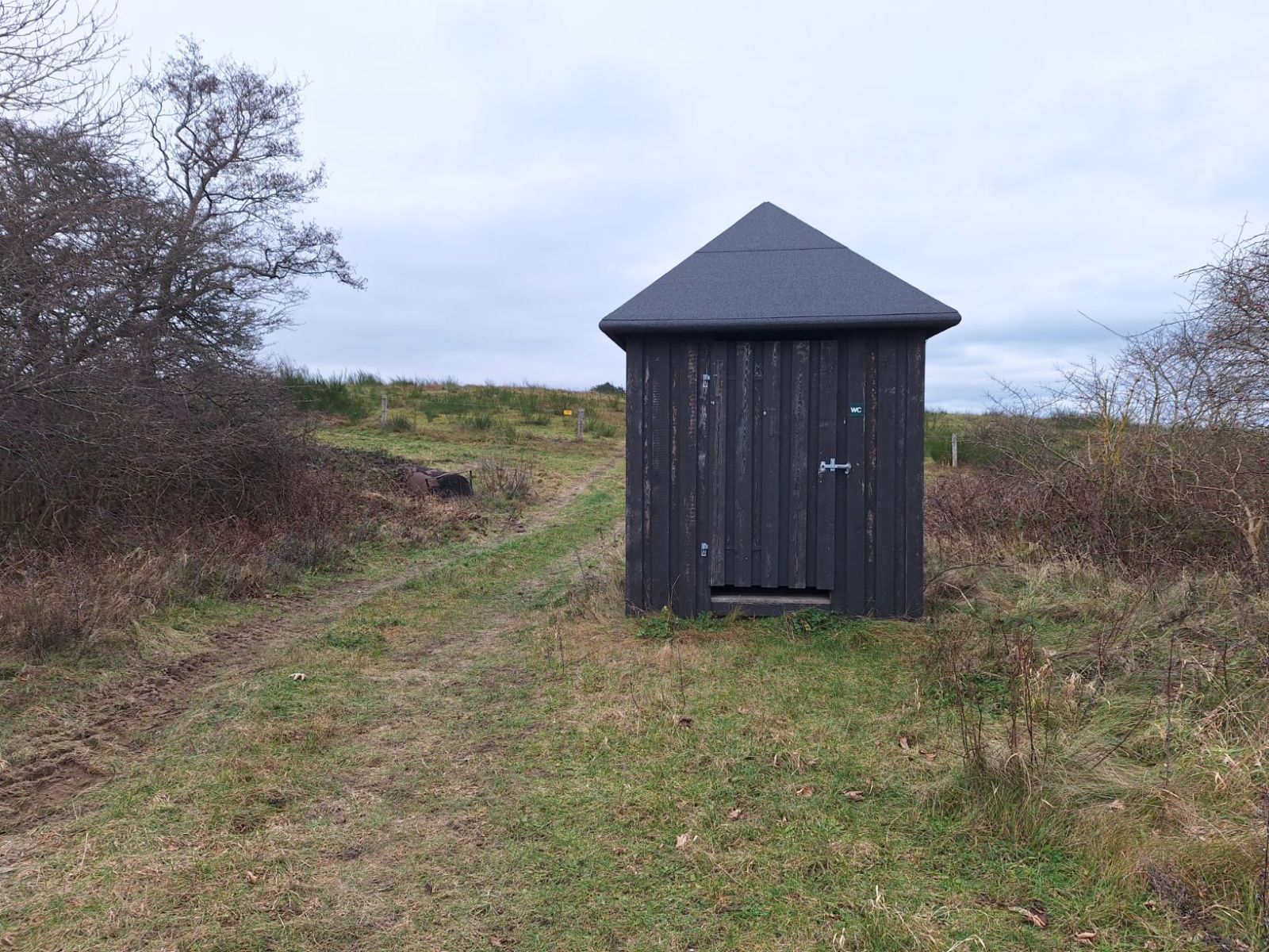 Toilettet ligger imellem to brede, levende hegn med en lille traktorpassage til venstre for. Indgangen til toilettet har et lille trin på omkring 10 centimeter. 